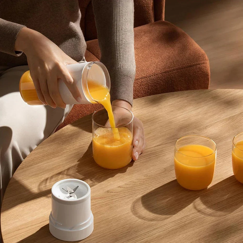 A person pours orange juice from a minimalist white portable blender into a glass on a wooden table.