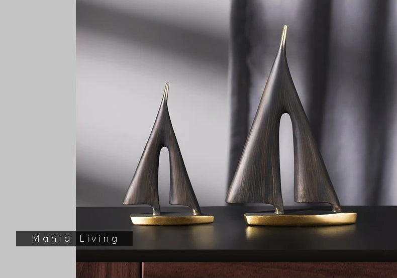Two minimalist, bronze-finish resin sailboat ornaments of different sizes on a dark surface.