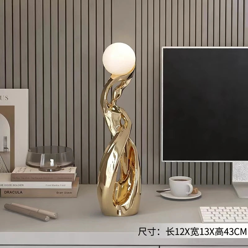 An abstract, gold-finished resin sculpture featuring a smooth, illuminated white sphere perched atop a twisted base.