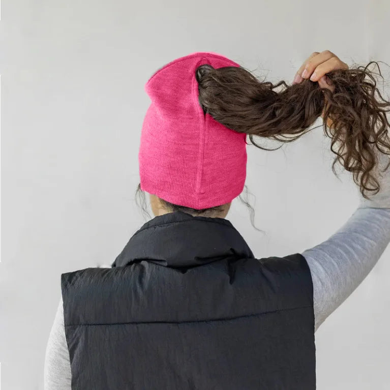 A woman wearing a vibrant pink knitted ponytail beanie, showing the back opening for hair, over a black gilet.