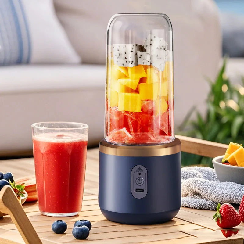 Portable electric blender in matte dark blue with gold trim, filled with cubed fruit, beside a glass of red juice.
