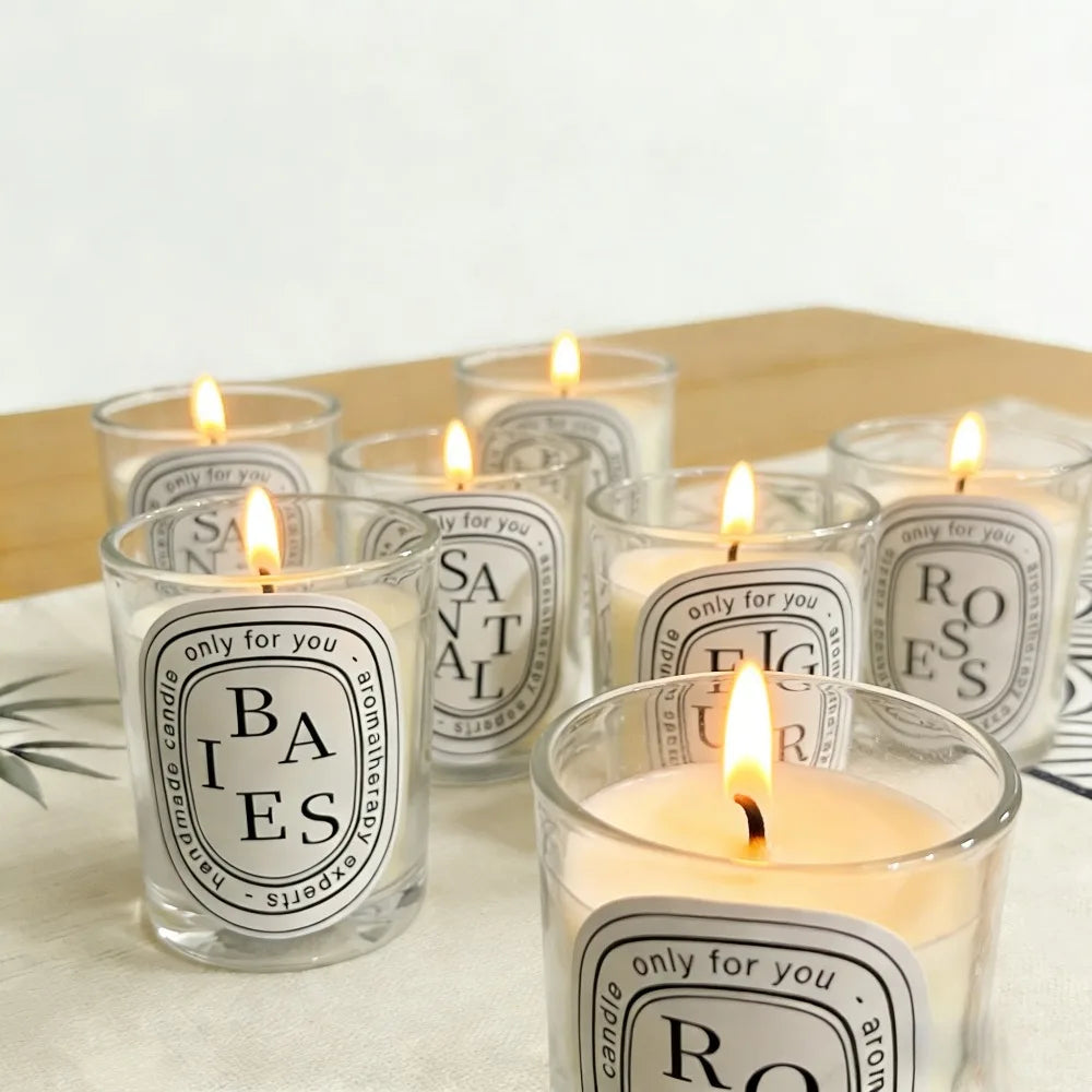 Seven lit aromatherapy candles in clear glass jars with oval labels, arranged on a white patterned surface.