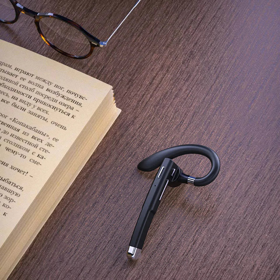 A sleek black wireless Bluetooth earpiece with a microphone hook, resting on a wooden desk next to a book and glasses.