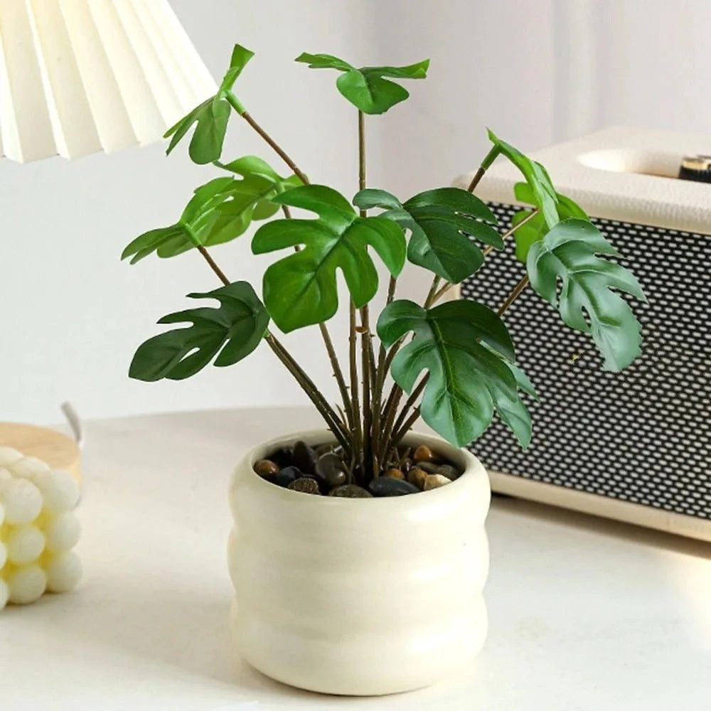 Artificial monstera plant in a cream, ribbed ceramic pot with river stones, set on a white table.