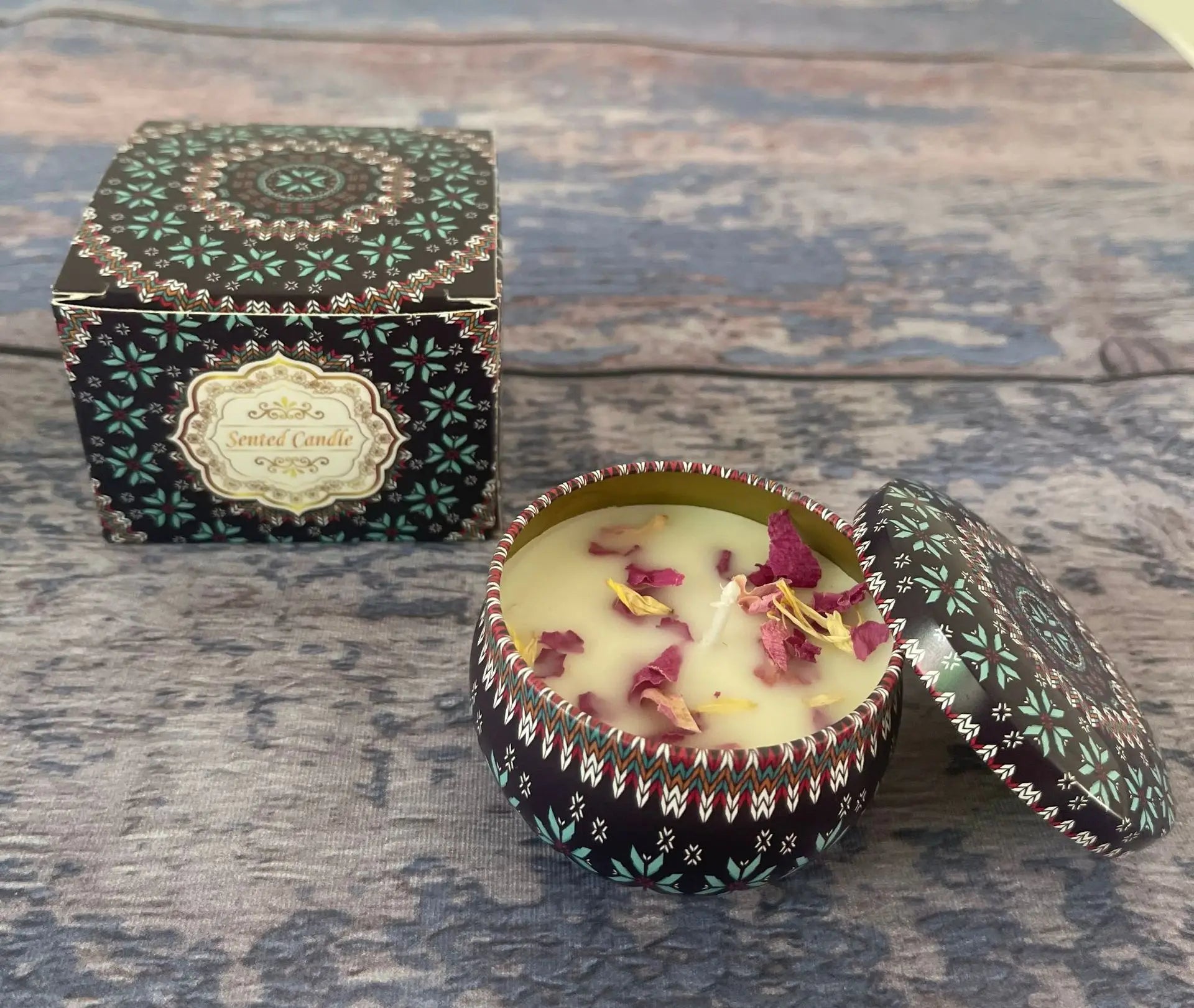 Scented candle in a decorated tin with a matching gift box, featuring floral petals and a dark patterned design.