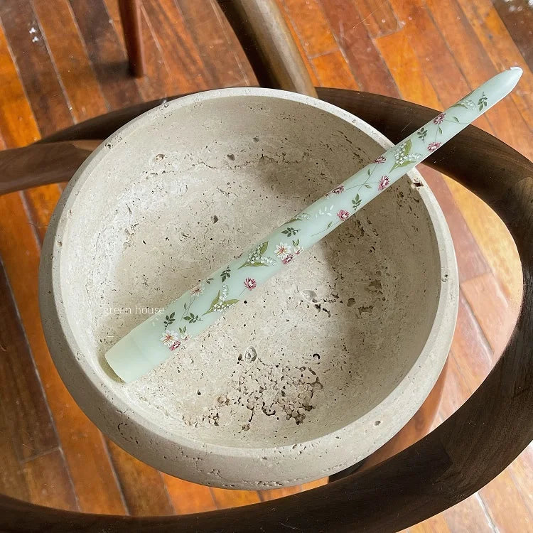 Pale green taper candle with a delicate floral print, resting inside a rustic, textured stone bowl.