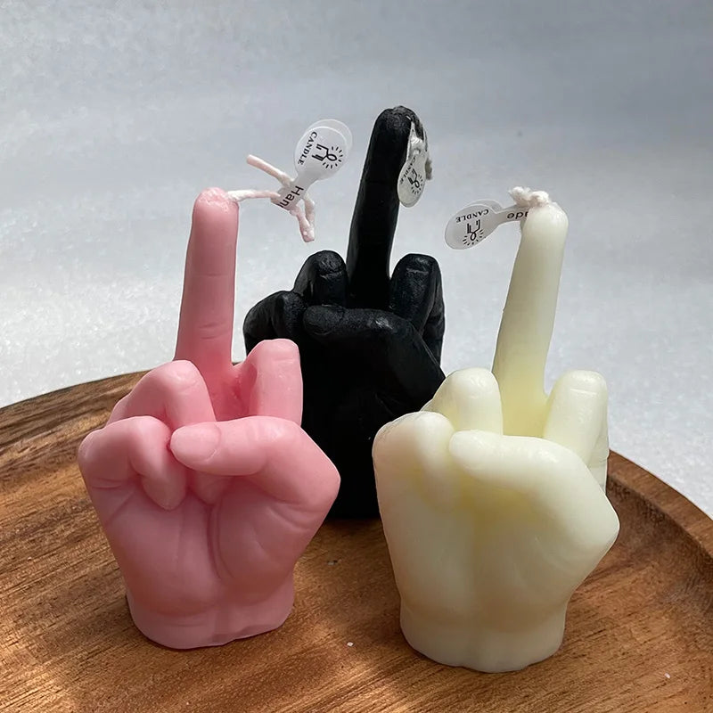 Three hand-shaped wax candles in pink, black, and white, each showing a middle finger gesture, on a wooden tray.