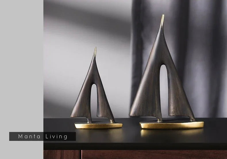 Two modern, minimalist sailboat ornaments in a dark bronze finish with gold-toned bases, displayed on a dark surface.