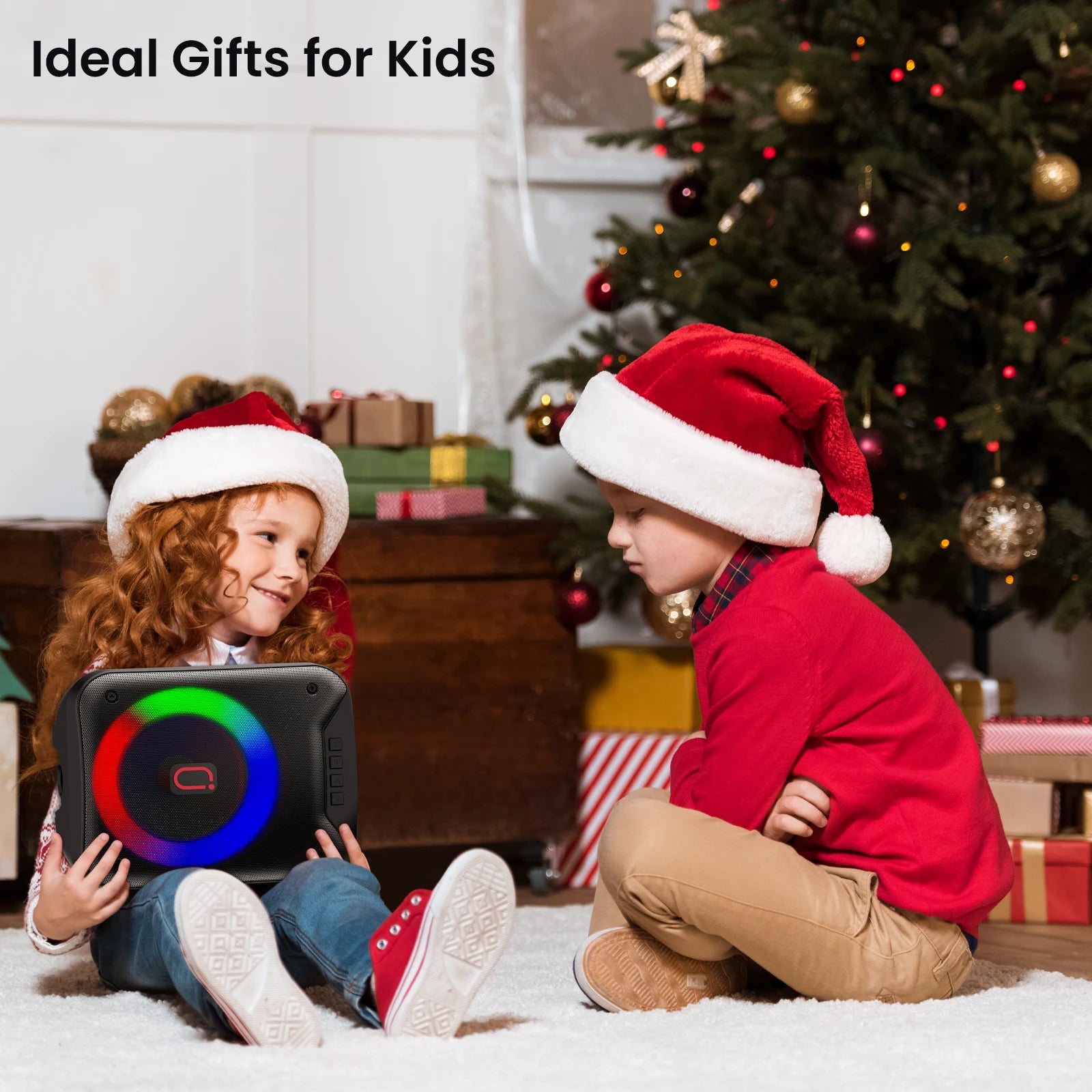 Two children in Santa hats sitting by a Christmas tree, holding a black portable wireless speaker with LED lights.