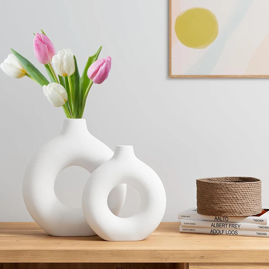 Two modern, white circular doughnut-shaped vases on a wooden surface, one holding a bouquet of pink and white tulips.
