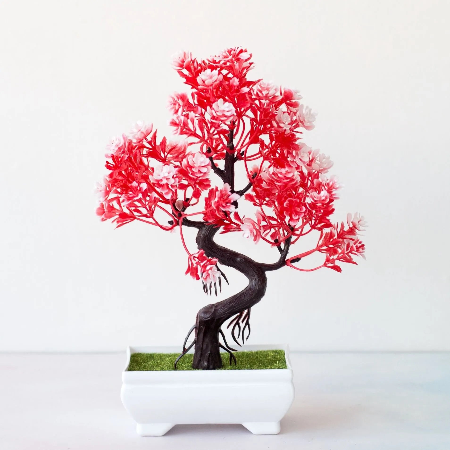 Artificial bonsai tree with vibrant red and white blossoms in a small white rectangular pot with green faux grass.