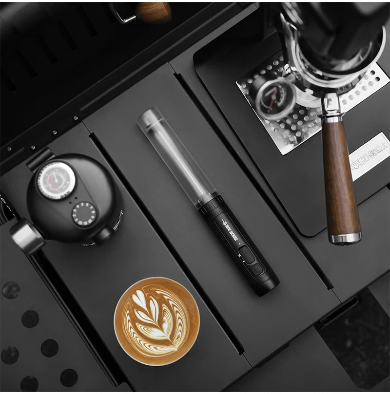 Black portable coffee frother wand on a matte black coffee station tray next to an espresso machine and latte art cup.