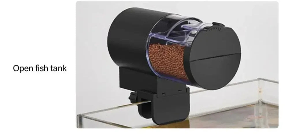Black automatic fish feeder with a clear food hopper attached to the edge of an open glass fish tank.