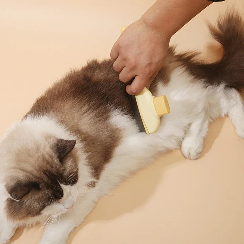 A person using a pale yellow grooming brush to groom a fluffy brown and white cat against a beige background.