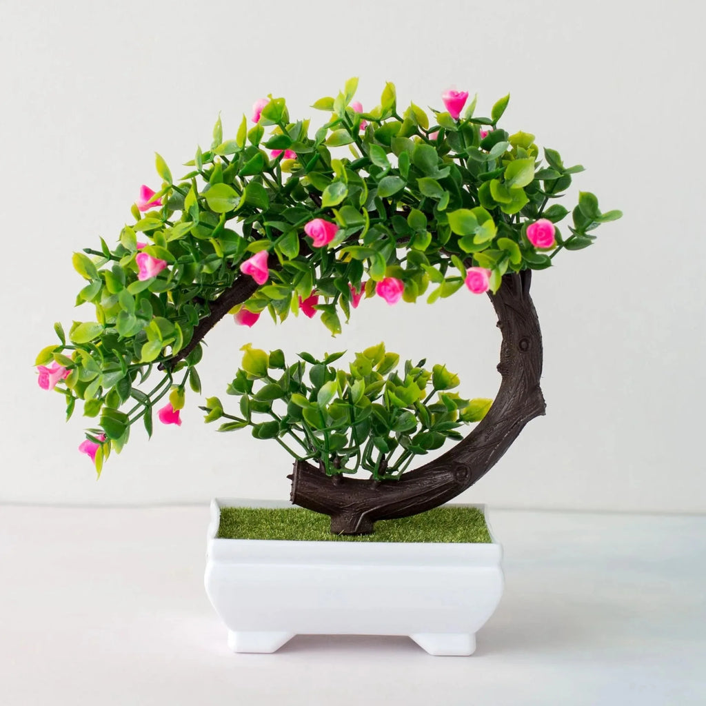Artificial bonsai with a curved brown trunk, green leaves, and small pink flowers in a white rectangular pot.