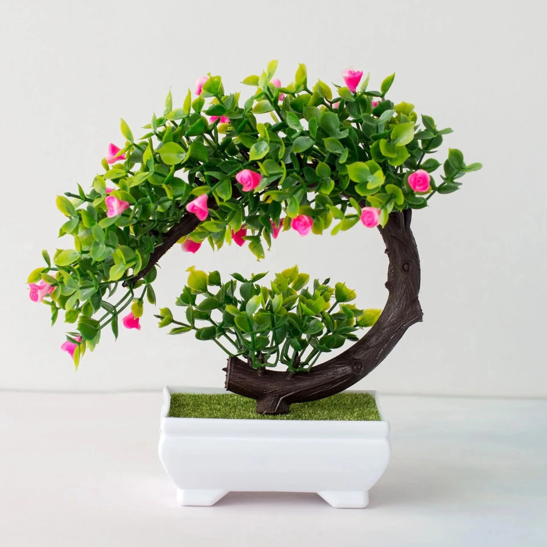 Artificial bonsai with a curved brown trunk, green leaves, and small pink flowers in a white rectangular pot.