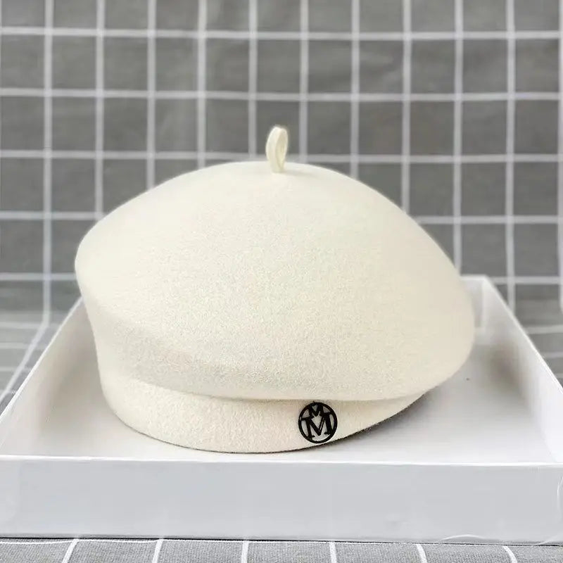 Classic cream wool felt beret with a small top loop and a circular black metallic detail on the brim.