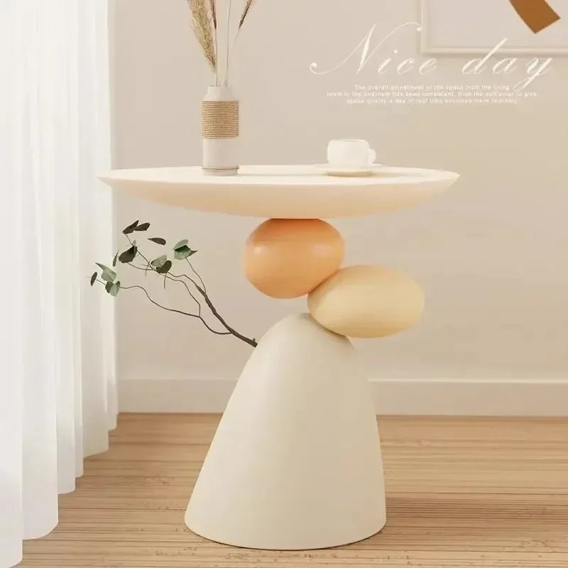 Minimalist cream side table featuring a round top supported by a unique, sculptural base of stacked pebble shapes.