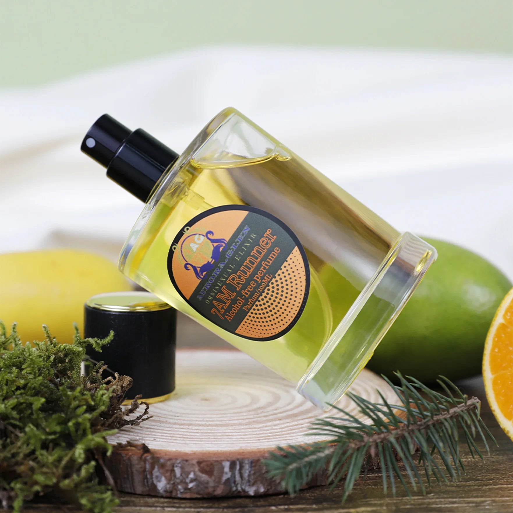 Clear glass spray bottle of alcohol-free yellow perfume on a wooden coaster with citrus fruits and greenery.