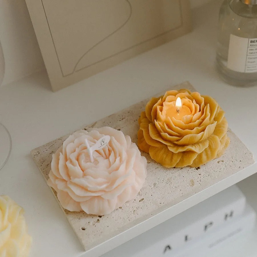 Two decorative flower-shaped candles, one soft pink and one yellow, on a textured stone tray. One candle is lit.