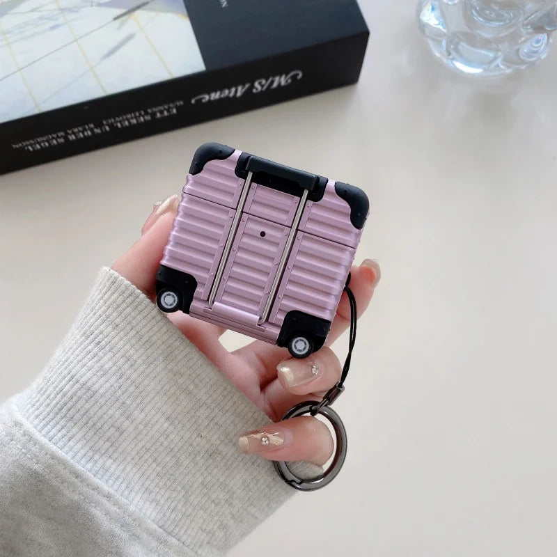 Pink suitcase-style hard shell AirPods case with keychain, featuring a ribbed design and black corner protectors.