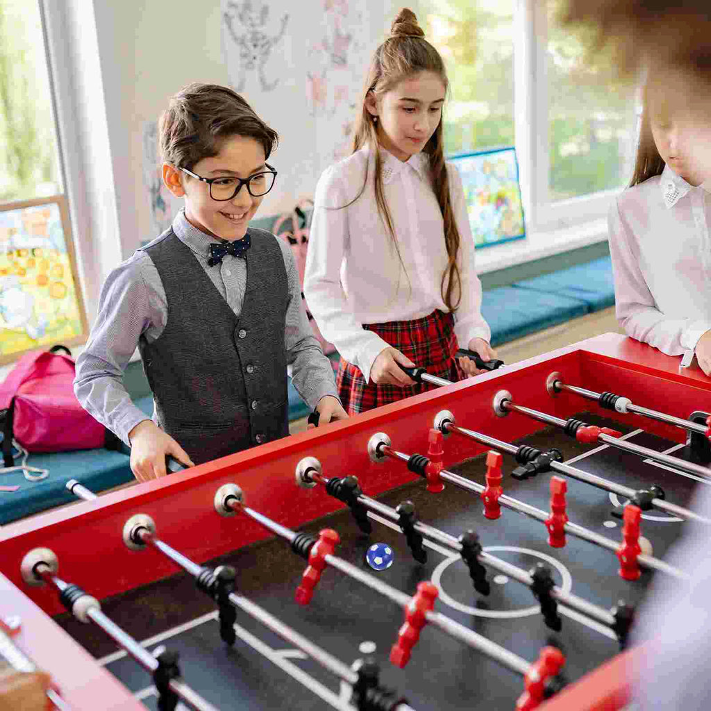 Children playing on a vibrant red foosball table with black pitch, metal rods, and red and black player figurines.