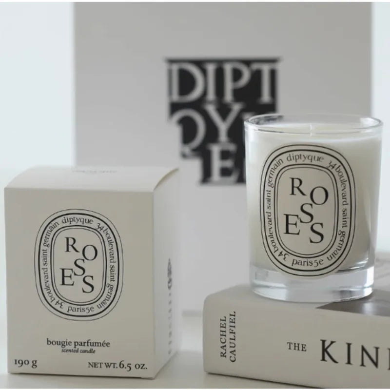 A scented candle in a clear glass jar with a white label, next to its matching cream-coloured box on a book.