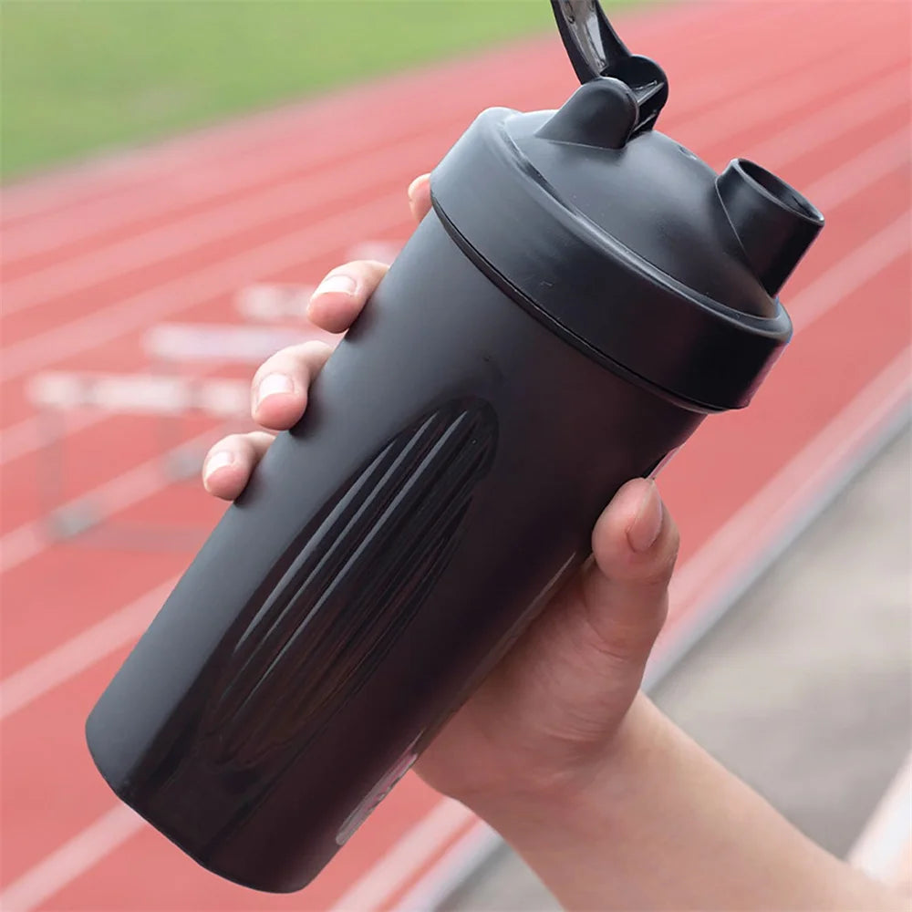 A hand holding a matte black protein shaker bottle with a flip-top lid, against a blurred athletics track background.