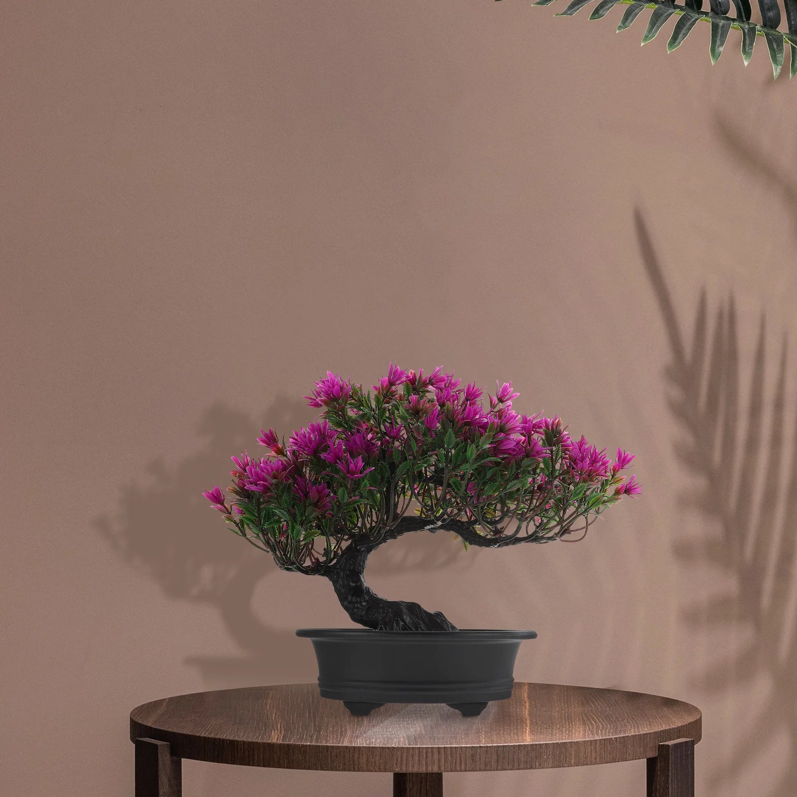 An artificial bonsai tree with vibrant magenta flowers in a black pot, displayed on a circular wooden table.
