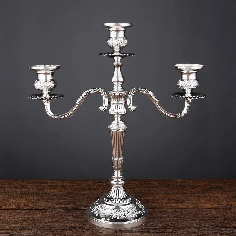 Silver metallic 3-arm candelabra with ornate floral embossing on the base and candle cups, standing on a wooden surface.