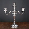 Silver metallic 3-arm candelabra with ornate floral embossing on the base and candle cups, standing on a wooden surface.