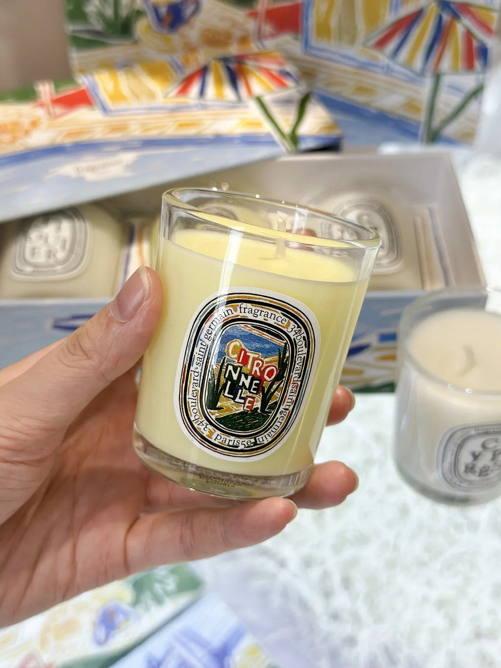 A hand holds a pale yellow scented candle in a clear glass jar with a colorful label featuring a landscape illustration.