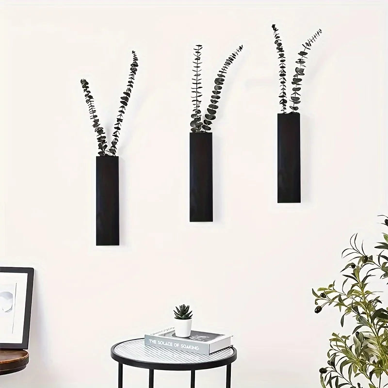 Three minimalist, black wooden wall-mounted vases holding dried eucalyptus stems displayed on a white wall.