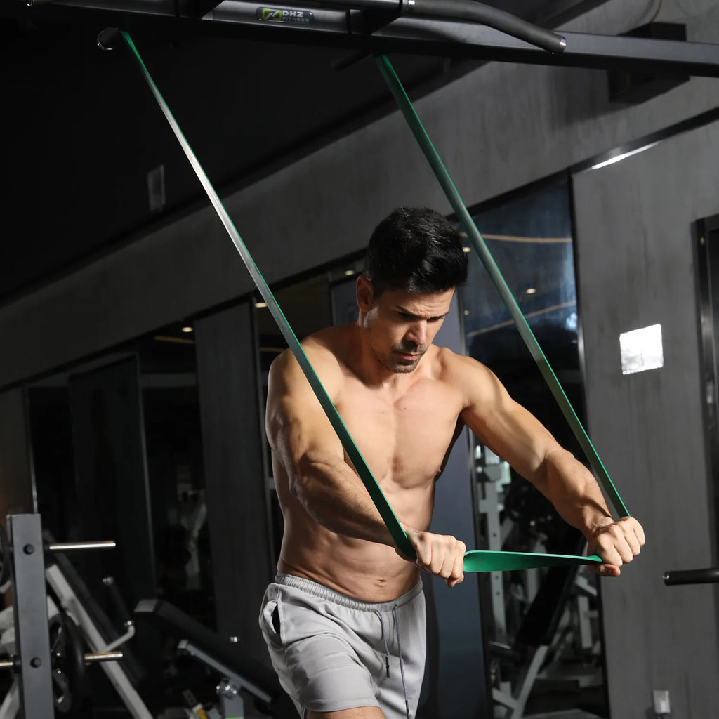 A man exercising in a gym using a long, green heavy-duty resistance band looped over a pull-up bar.