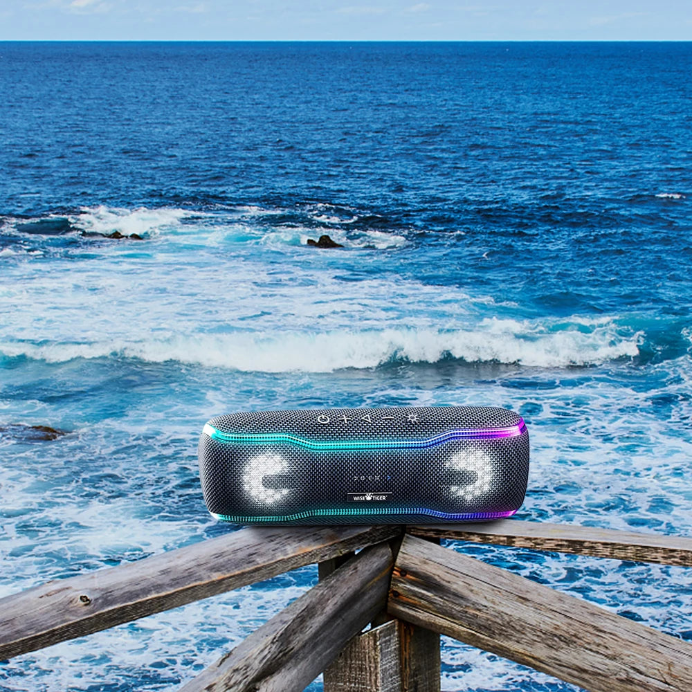 Portable black Bluetooth speaker with vibrant LED light strips, resting on a wooden railing overlooking the sea.