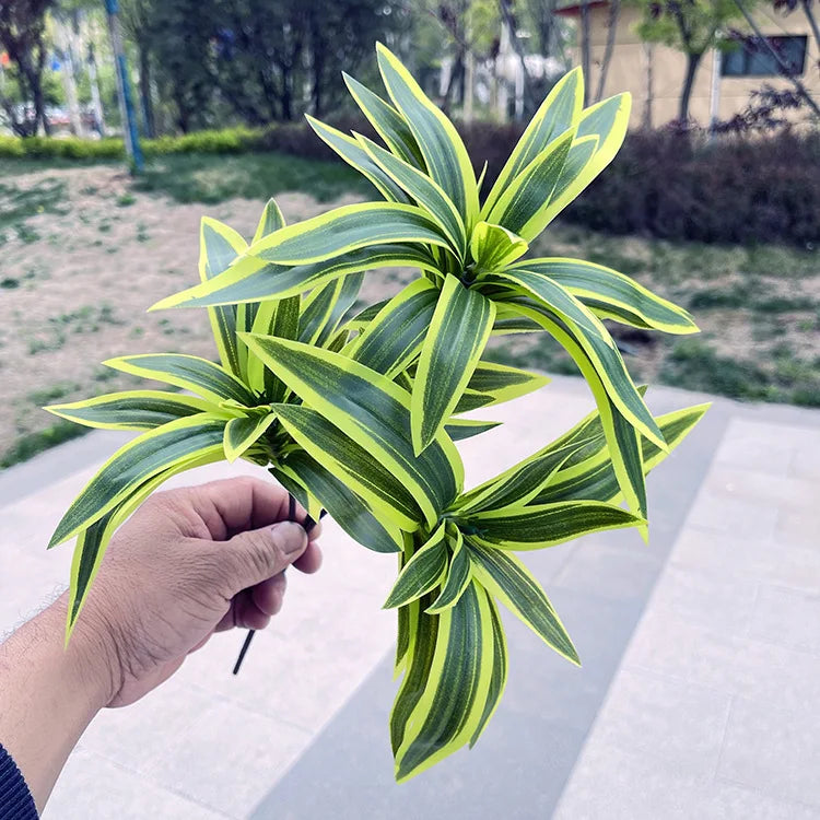 A hand holding an artificial plant branch with pointed, variegated lime green and dark green striped leaves.