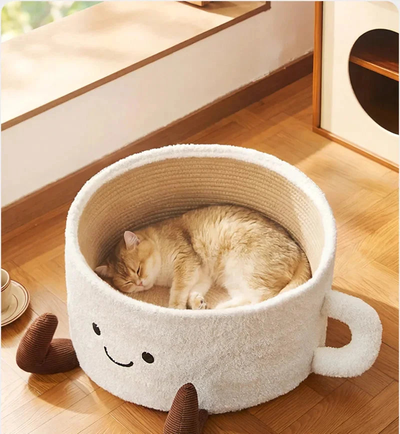 White coffee cup-shaped cat bed with smiling face, handle, and brown feet, holding a sleeping cat.