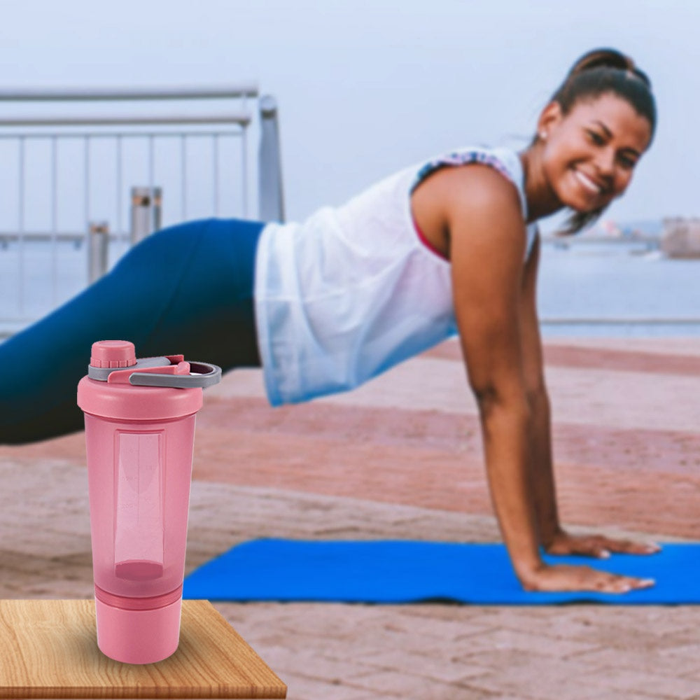 Pink plastic shaker bottle with a grey lid and storage compartment, set against a blurred background of a woman in plank.
