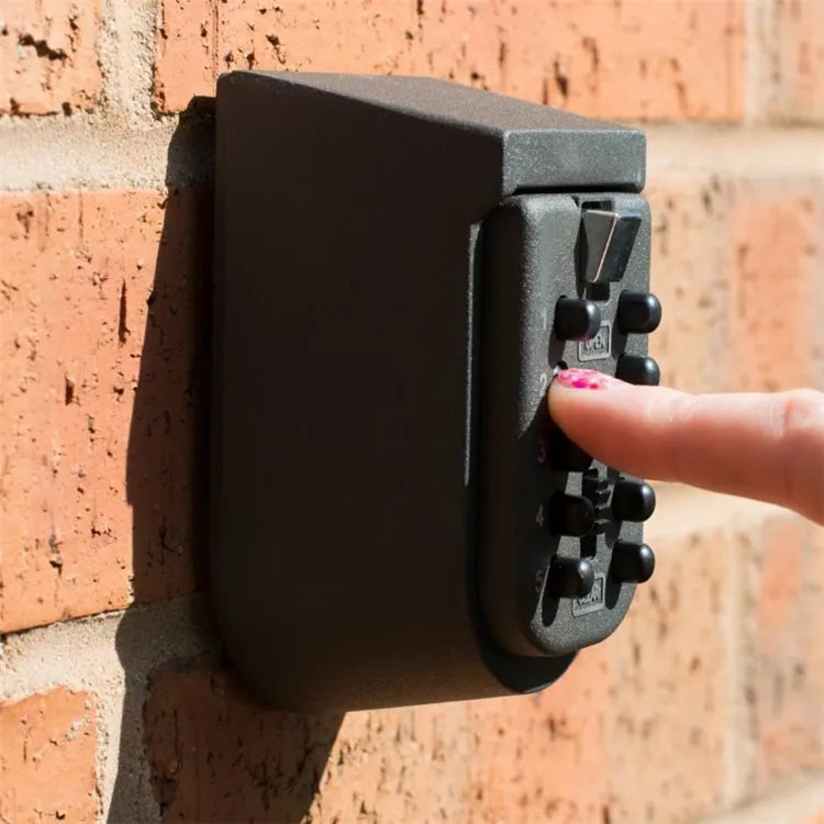 Dark gray wall-mounted key lock box with push-button combination, being pressed by a finger on a brick wall.