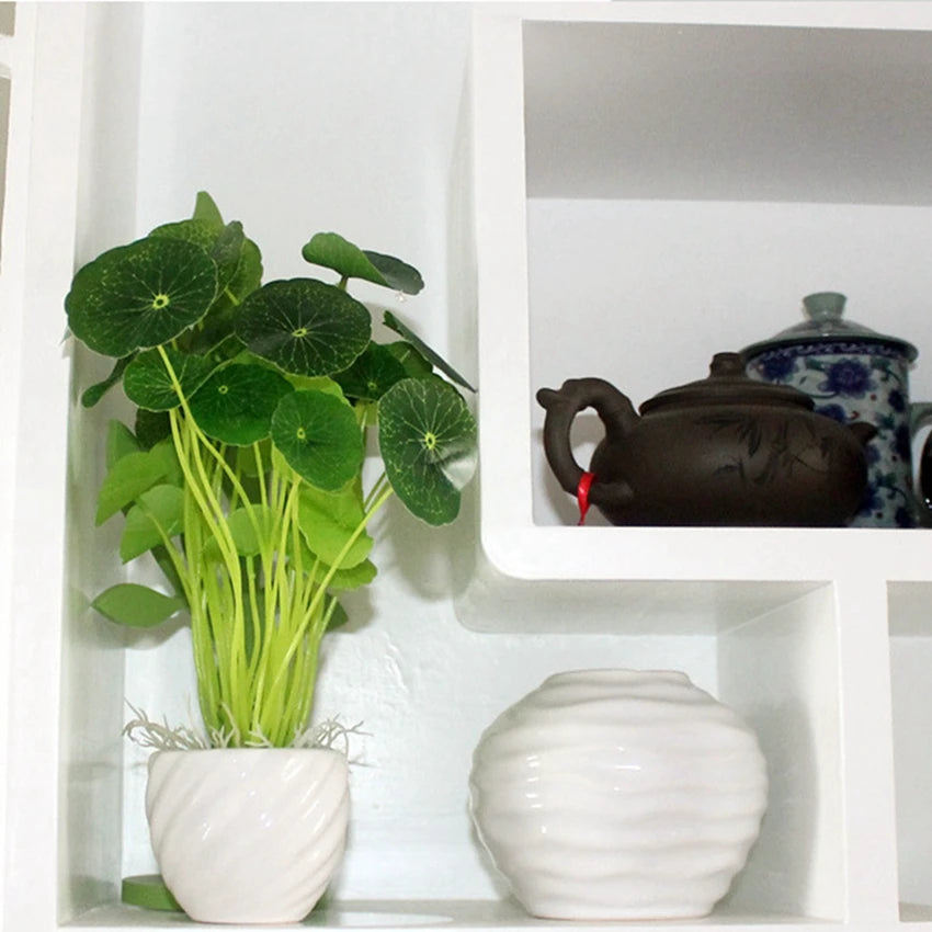Artificial lotus plant with round green leaves in a white swirled pot, displayed on a white shelf next to decor.