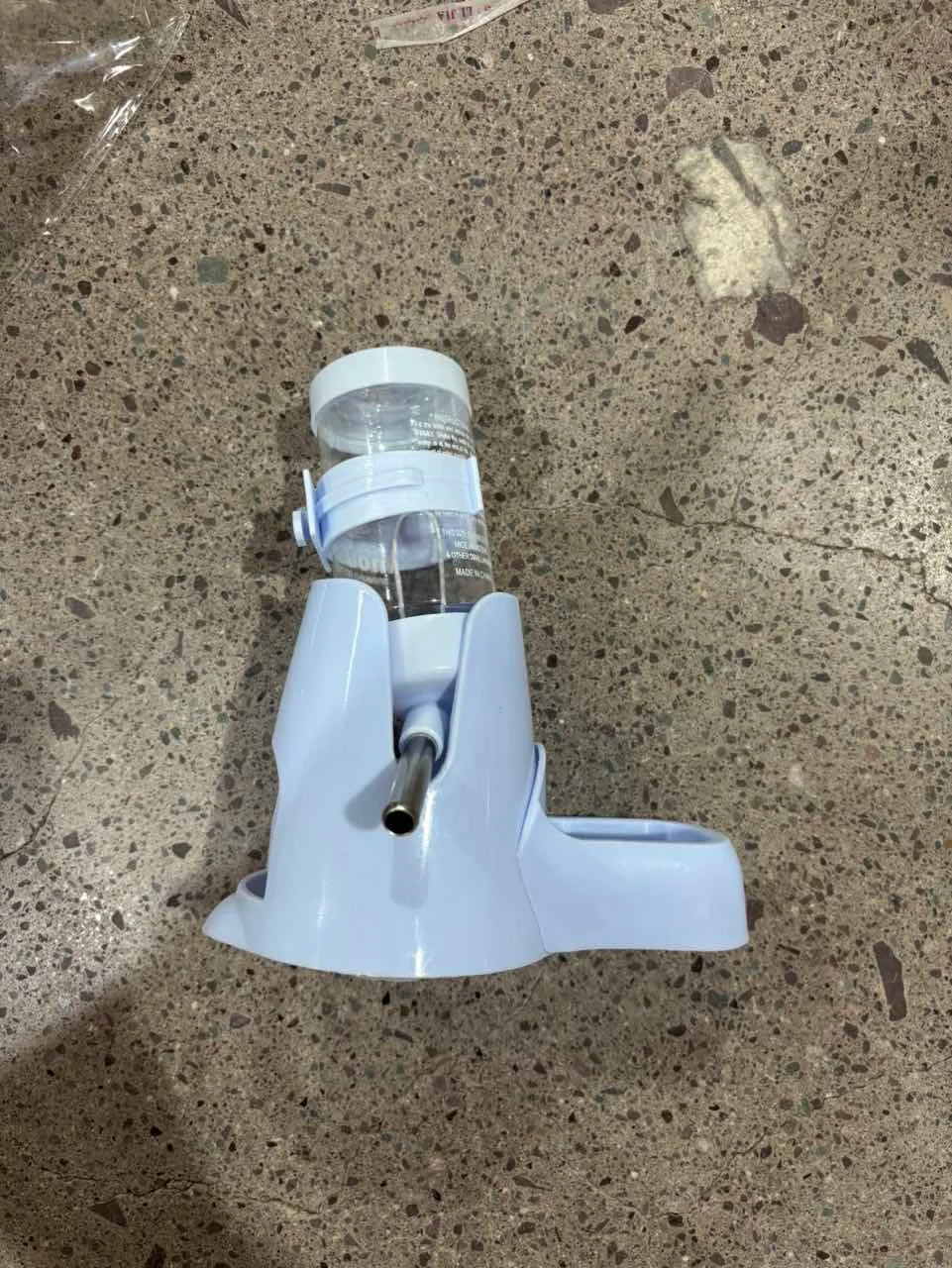 Light blue pet water dispenser with a clear plastic bottle and metal nozzle on a grey speckled floor.