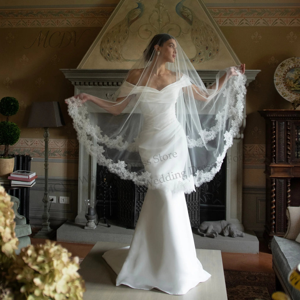 White off-the-shoulder mermaid satin wedding dress with sweetheart neckline and a sheer lace-trim veil.