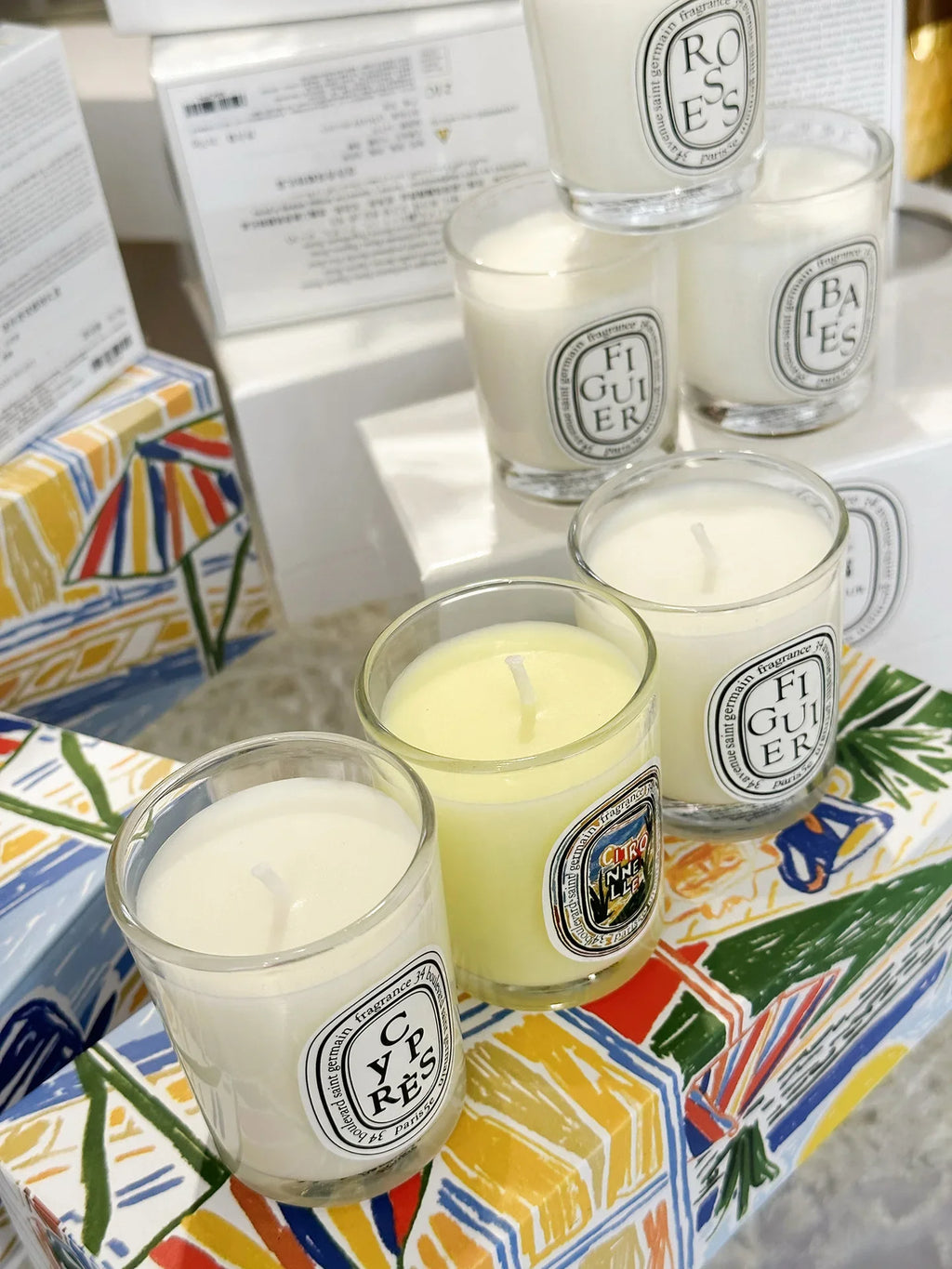 Collection of scented candles in clear glass jars with oval labels, arranged on colourful patterned boxes.