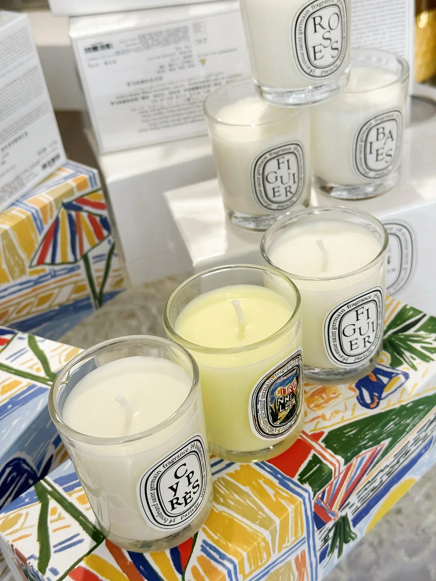 Collection of scented candles in clear glass jars with oval labels, arranged on colourful patterned boxes.