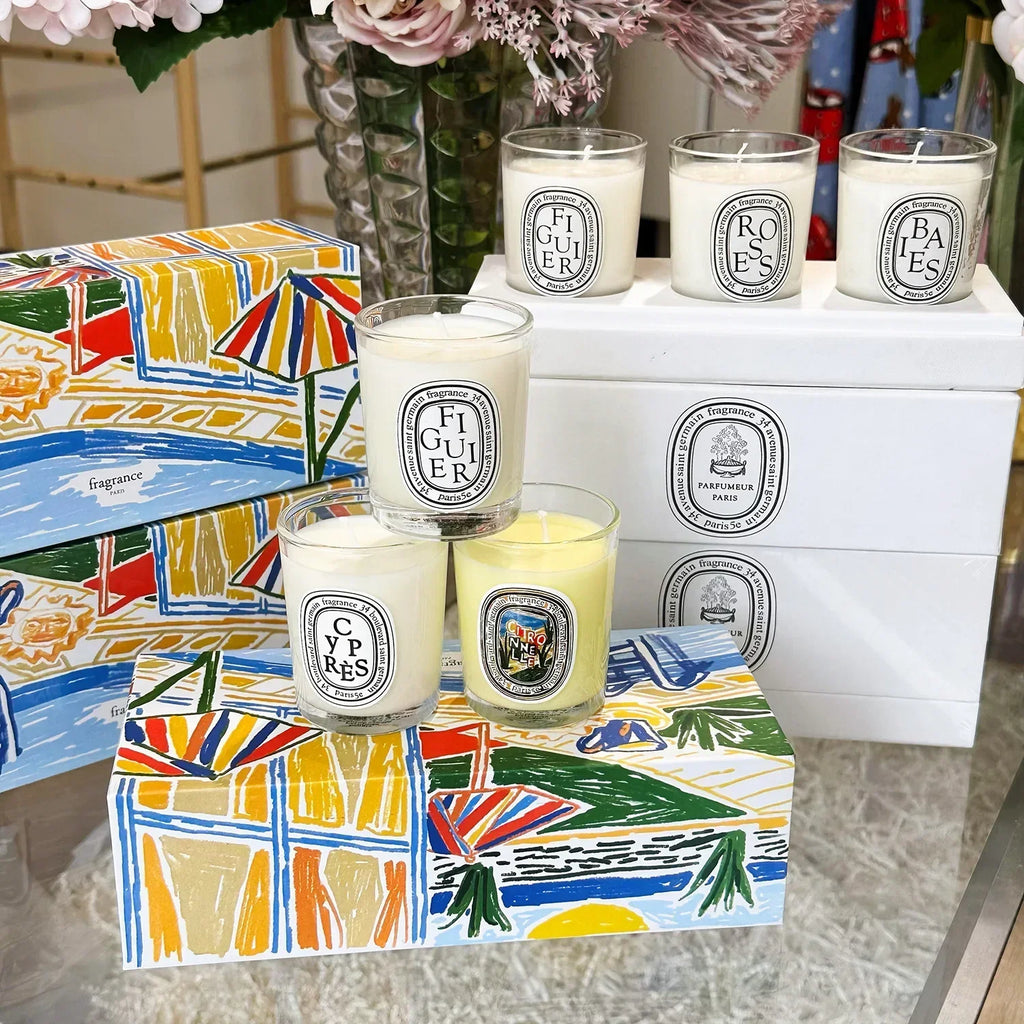 Set of scented candles in clear glass jars with oval labels, presented in decorative boxes featuring vibrant illustrations.