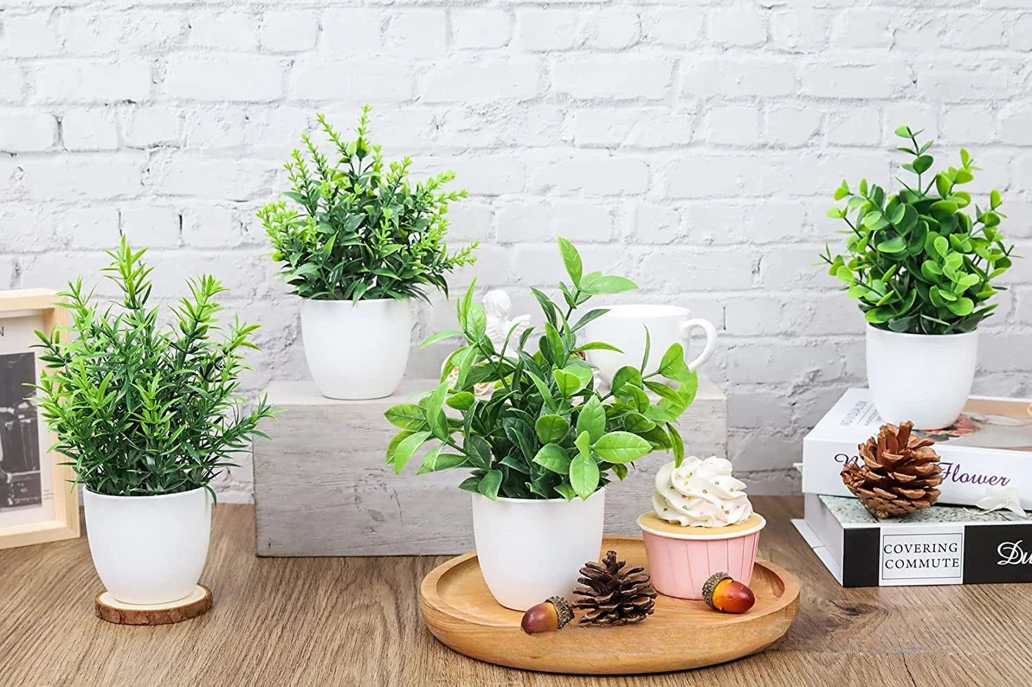 Four artificial green potted plants in small white pots, arranged on a wooden table with decorative accents.