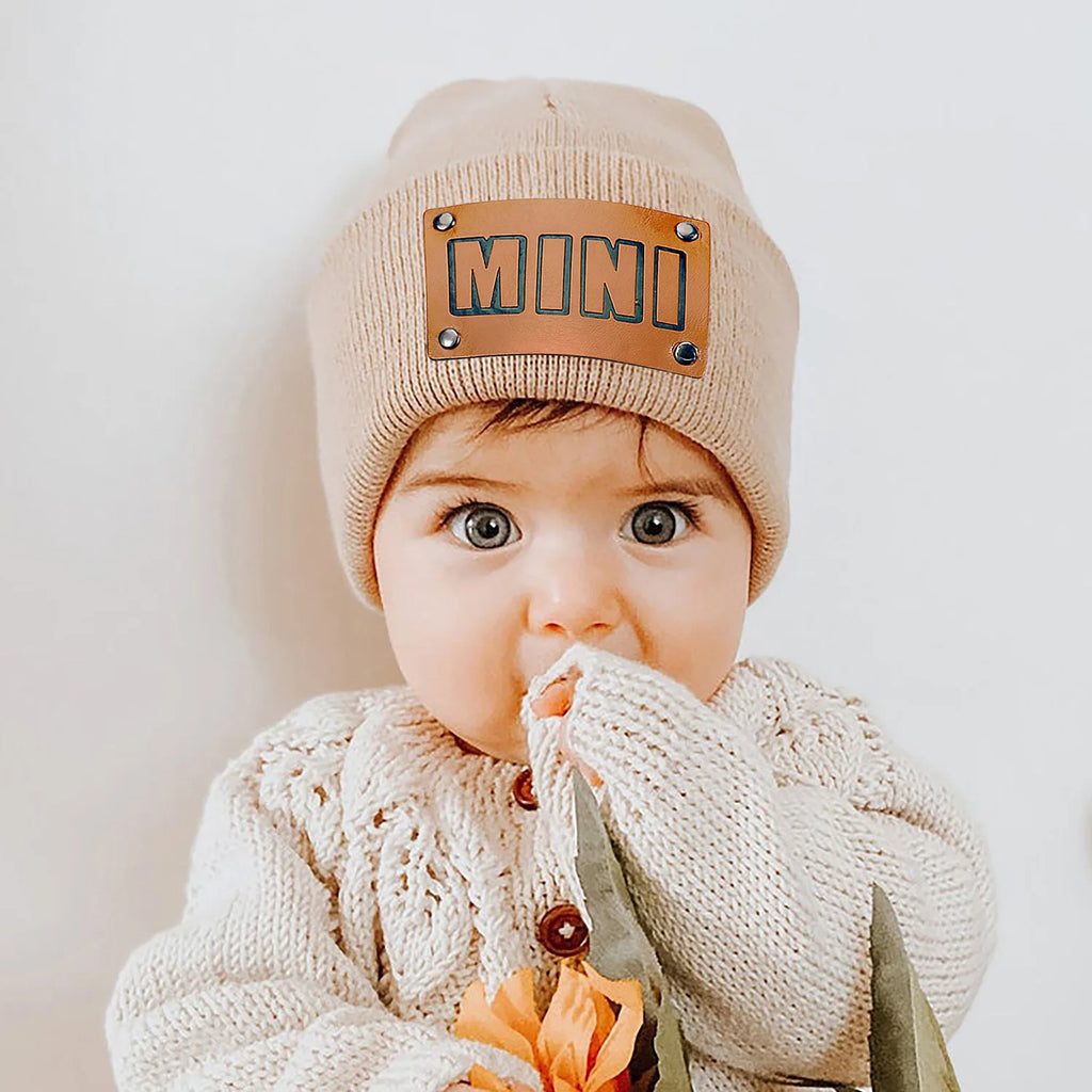 A baby wearing a beige ribbed beanie with a brown leather-look patch featuring the word 'MINI' and stud details.