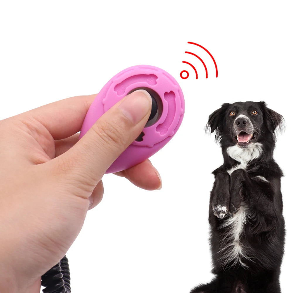 A hand holding a pink, ergonomic dog training clicker with a wrist coil, next to an attentive black and white dog.