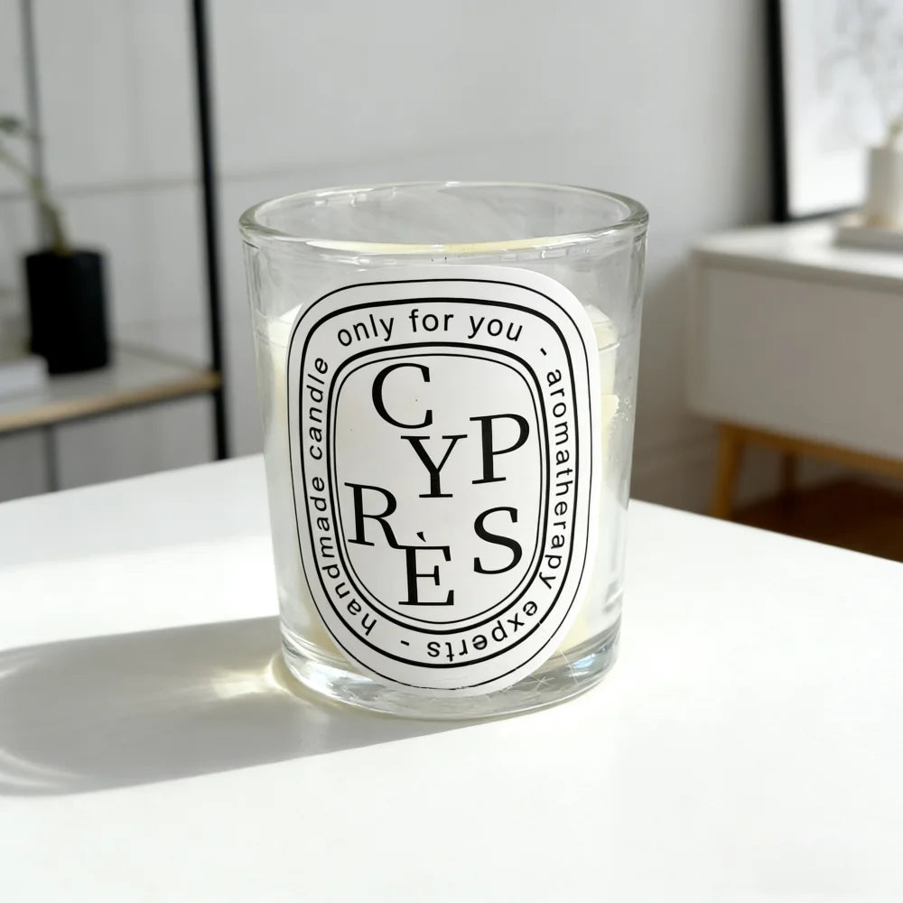 A scented candle in a clear glass jar with a white oval label reading "CYPRES" and black typography on a white table.