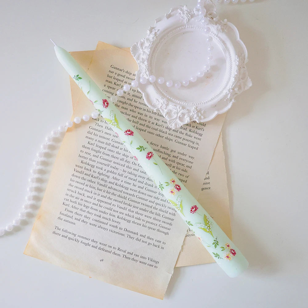 Pale green tapered candle decorated with delicate floral patterns, resting on vintage book pages with pearl accents.